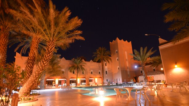 A nighttime view of a courtyard with a pool, palm trees, and a building with multiple balconies. - Ksar Tinsouline (Zagora)