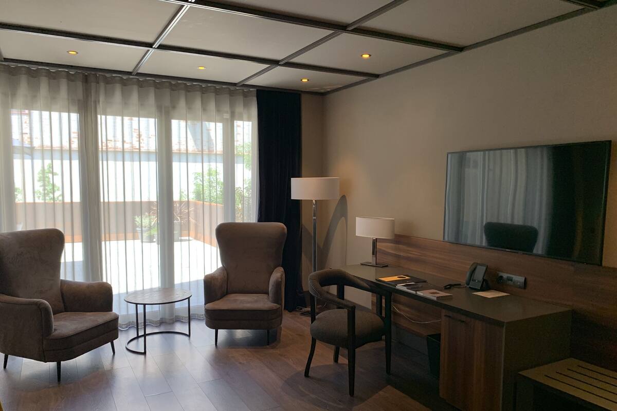 Habitación de hotel con escritorio, dos sillones, una mesita, lámpara y un ventanal grande con cortinas.