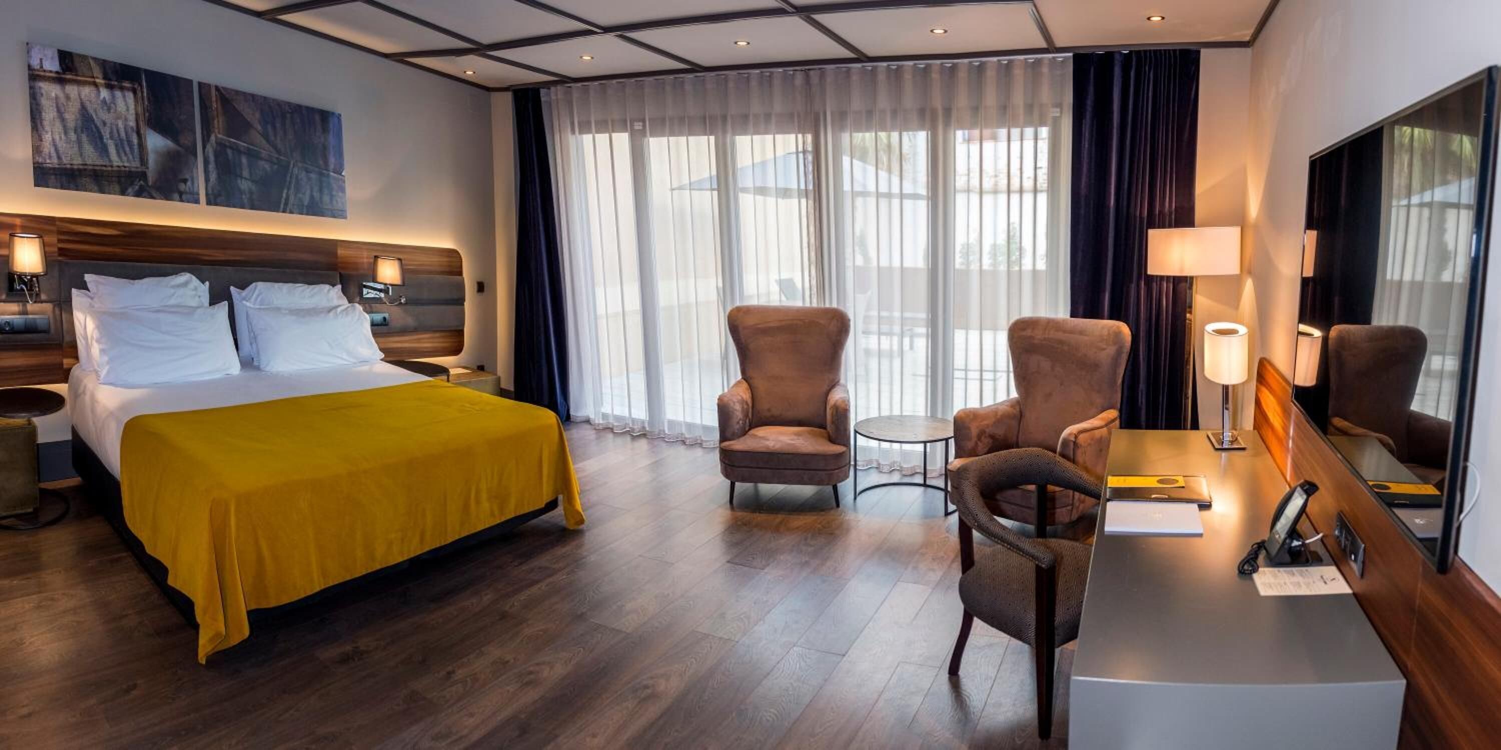 A hotel room with a bed, two armchairs, a desk with a lamp, and a large mirror.