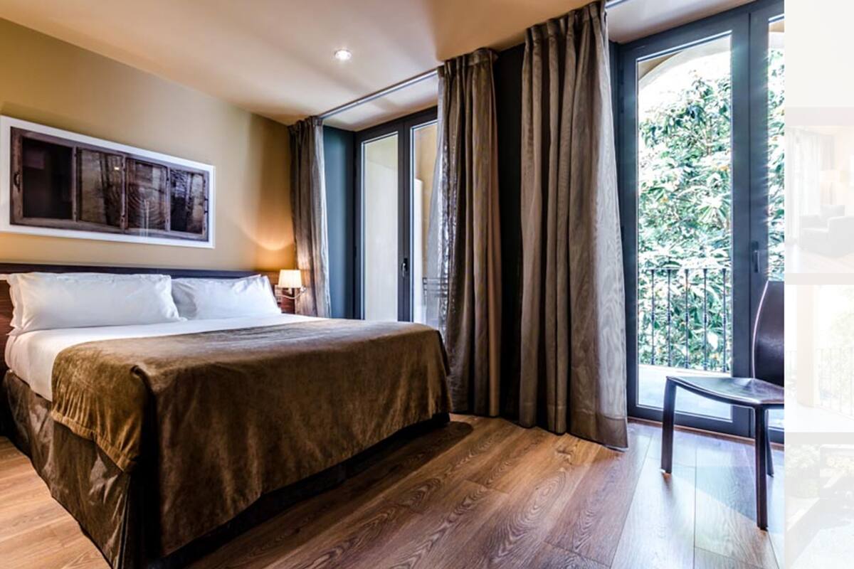 Habitación de hotel moderna con una cama grande, vista a zonas verdes por la ventana y una silla junto a ella.