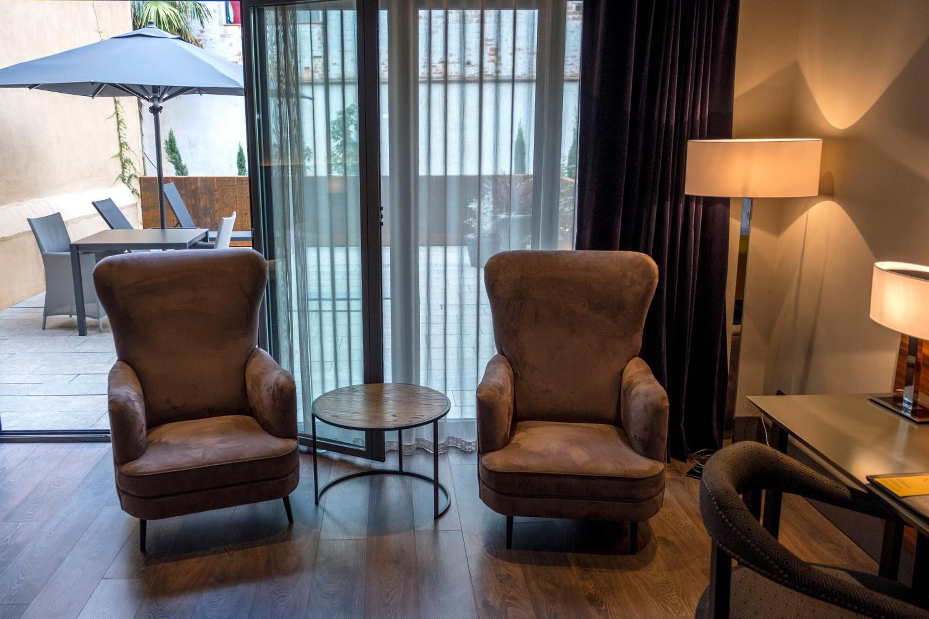 A hotel room with two armchairs, a small round table, a lamp, and a view through a glass door.