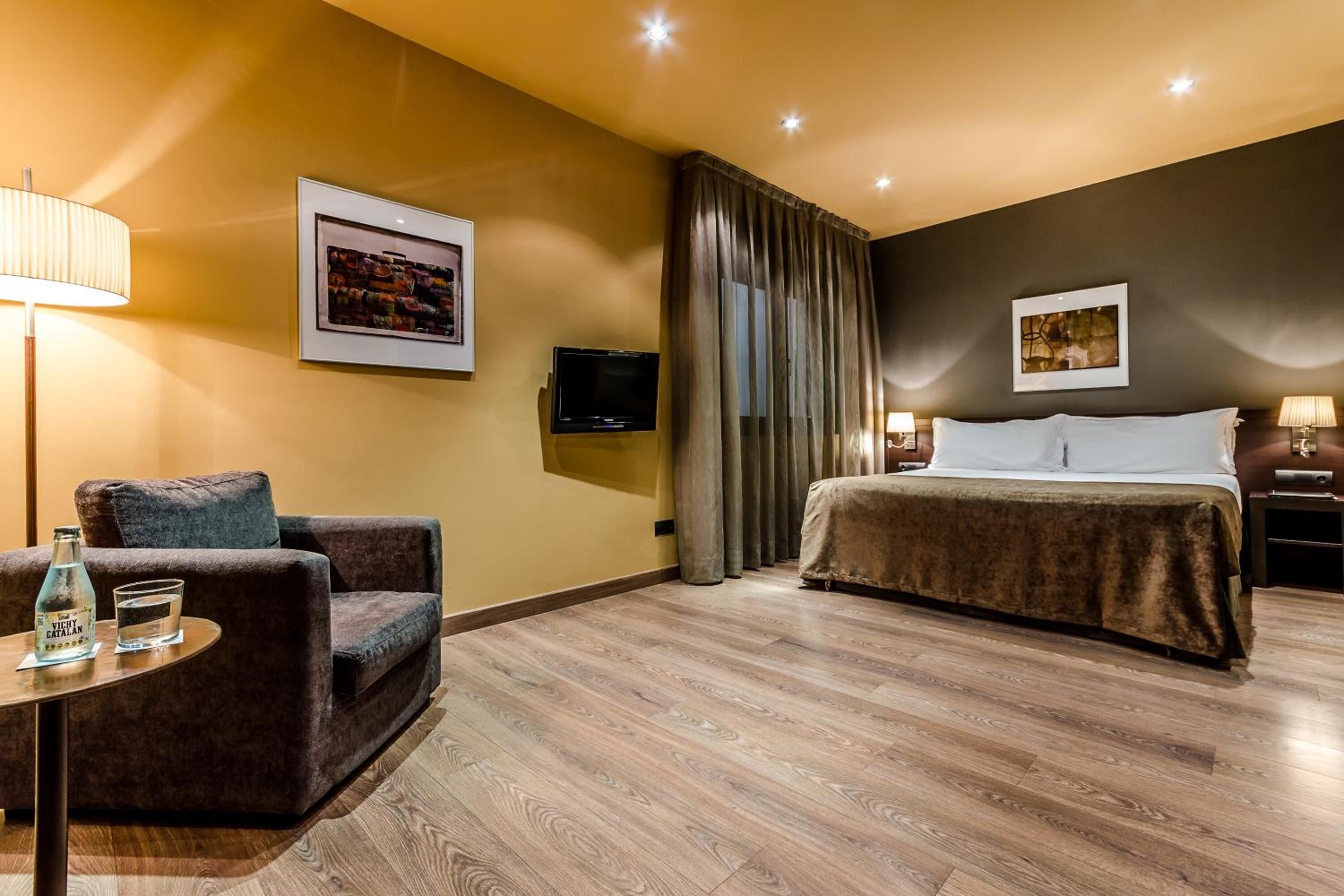 A hotel room with a bed, a sofa, a TV, and a table with a bottle and glass.