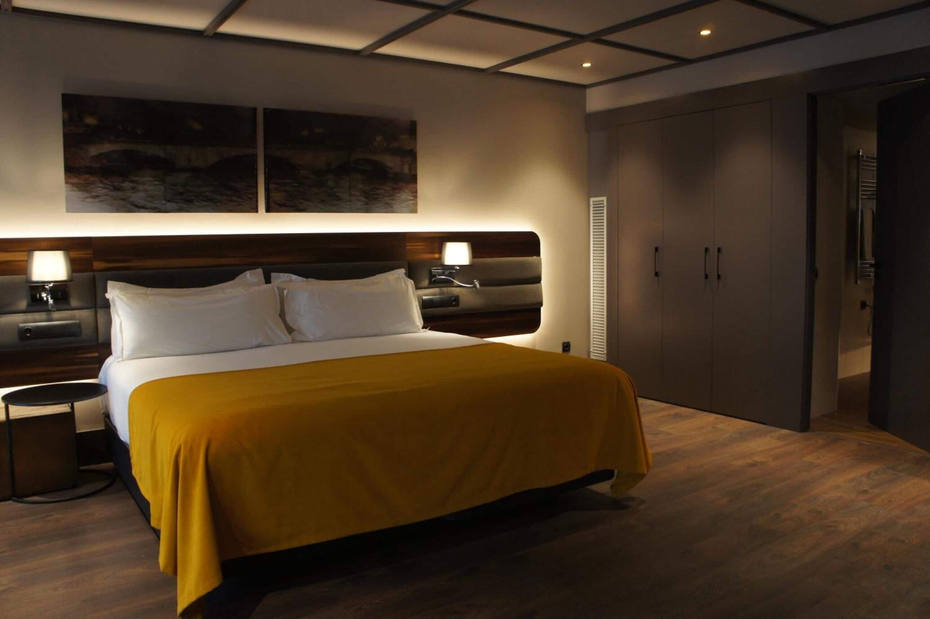 A modern hotel room with a large bed, bedside tables, a headboard with integrated lighting, and a wardrobe.
