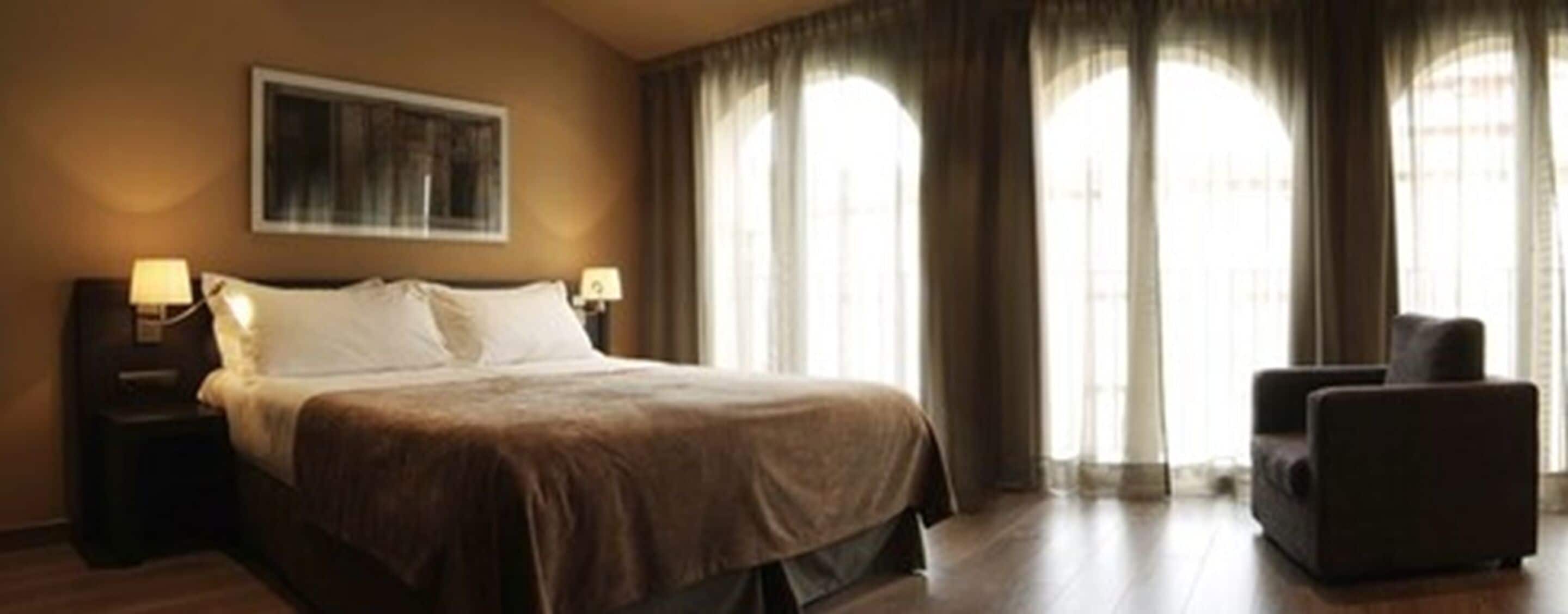Una habitación de hotel con una cama, dos lámparas de noche, un sillón y ventanas amplias con cortinas.