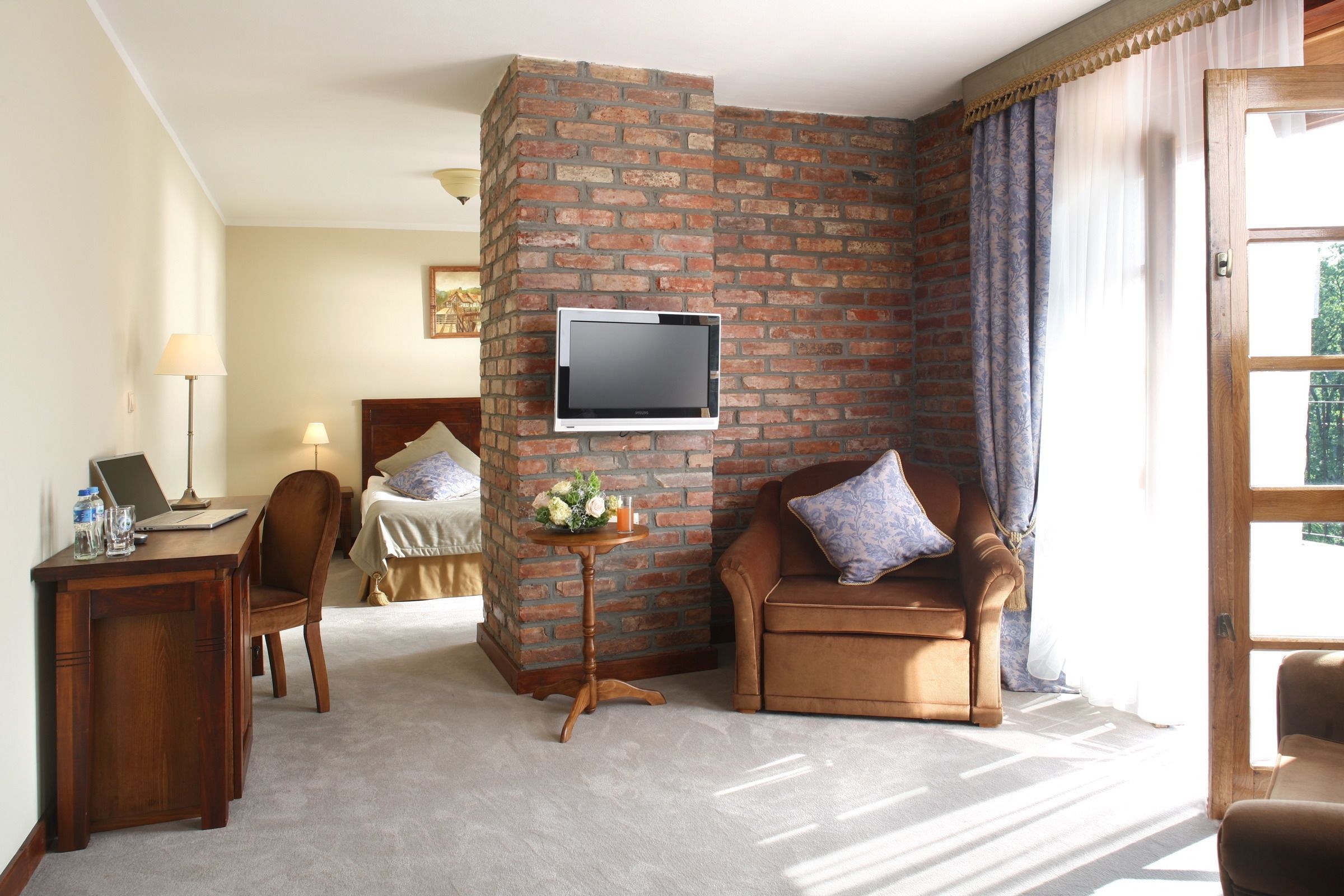 A hotel room with a brick accent wall, a flat-screen TV mounted above a wooden desk, a brown leather armchair, a small round table with a vase of flowers, and a bed with a white headboard and pillows.