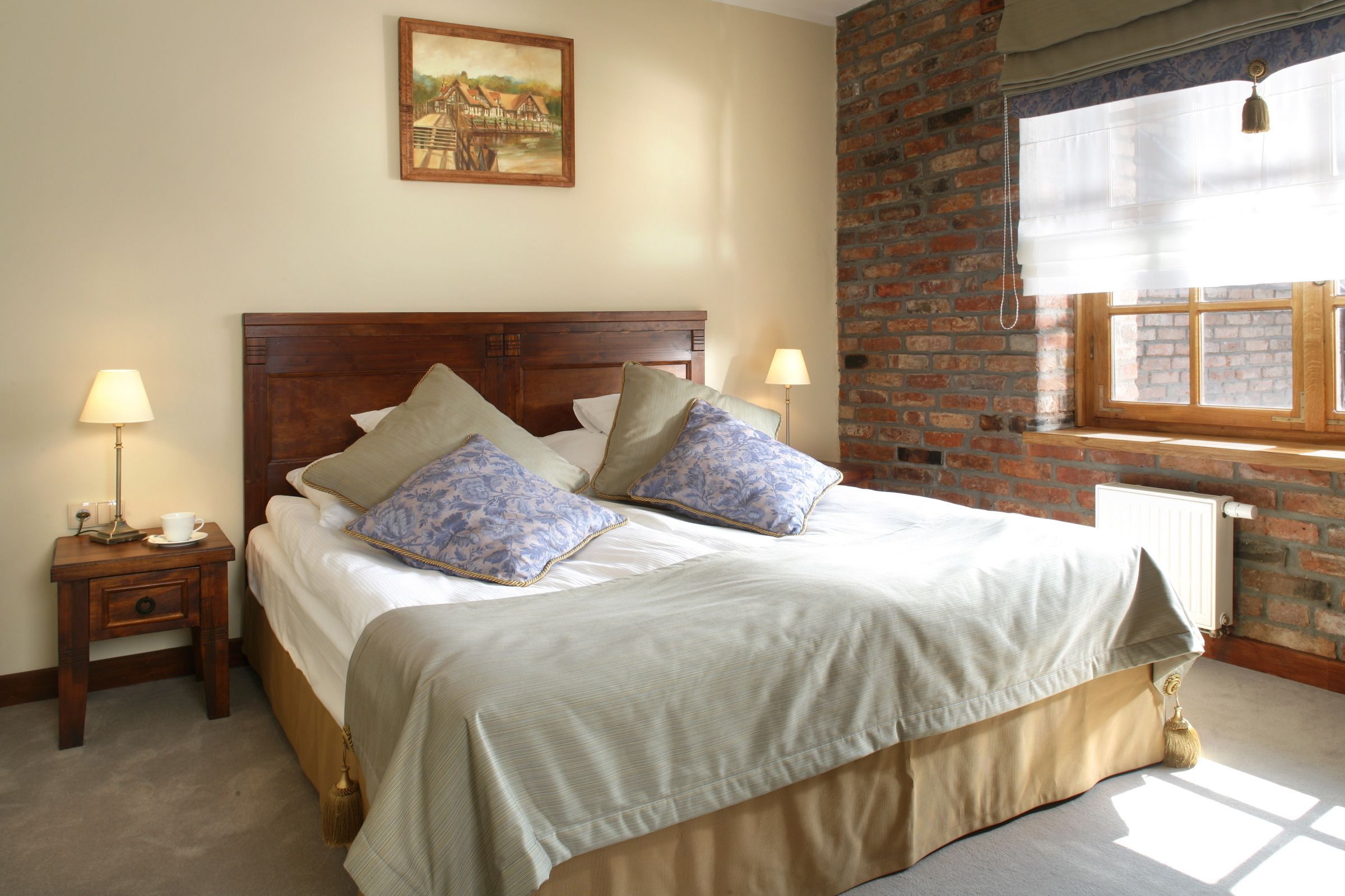 Uma cama bem arrumada com cabeceira de madeira, duas mesas de cabeceira com abajures, um quadro na parede e uma parede de tijolos ao lado de uma janela com persiana branca.