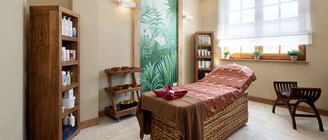 Un dormitorio con cama, una estantería de madera, repisas en la pared con artículos de tocador, una ventana con plantas y una silla.