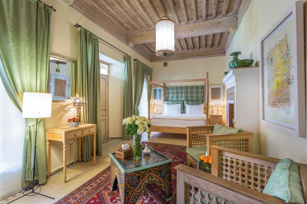 Quarto tradicional com uma cama, área de estar com mesa e cadeiras, um espelho e uma cortina verde.