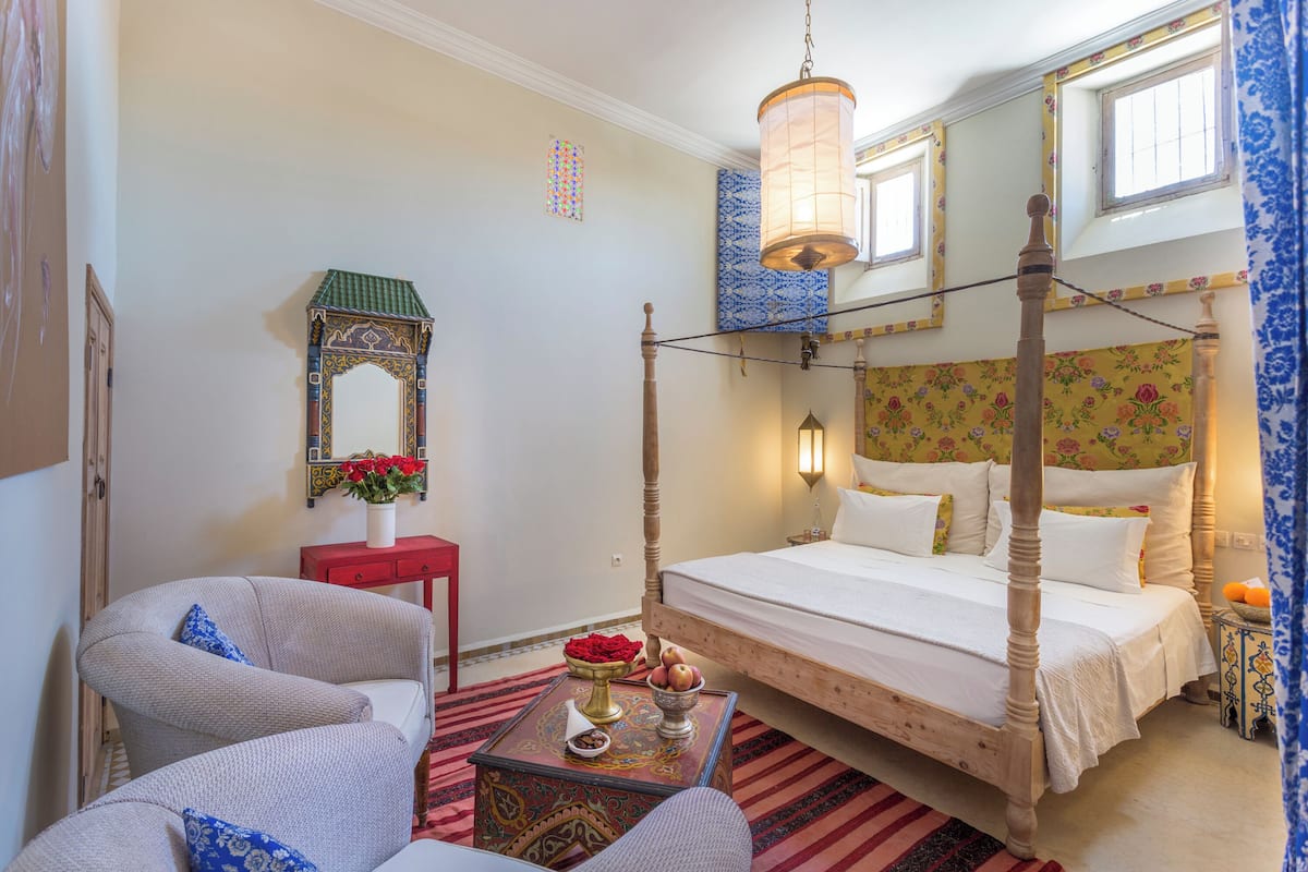 Uma cama com dossel e estrutura de madeira, uma mesa de cabeceira vermelha com uma tigela de frutas e uma cadeira com almofada azul.