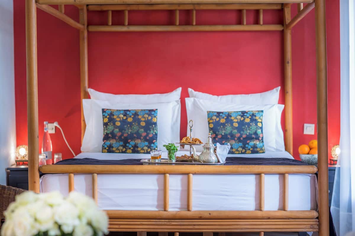 Uma cama com estrutura de madeira, roupa de cama branca e almofadas decorativas. Um tabuleiro com bule, copos e uma pequena planta está sobre a cama.