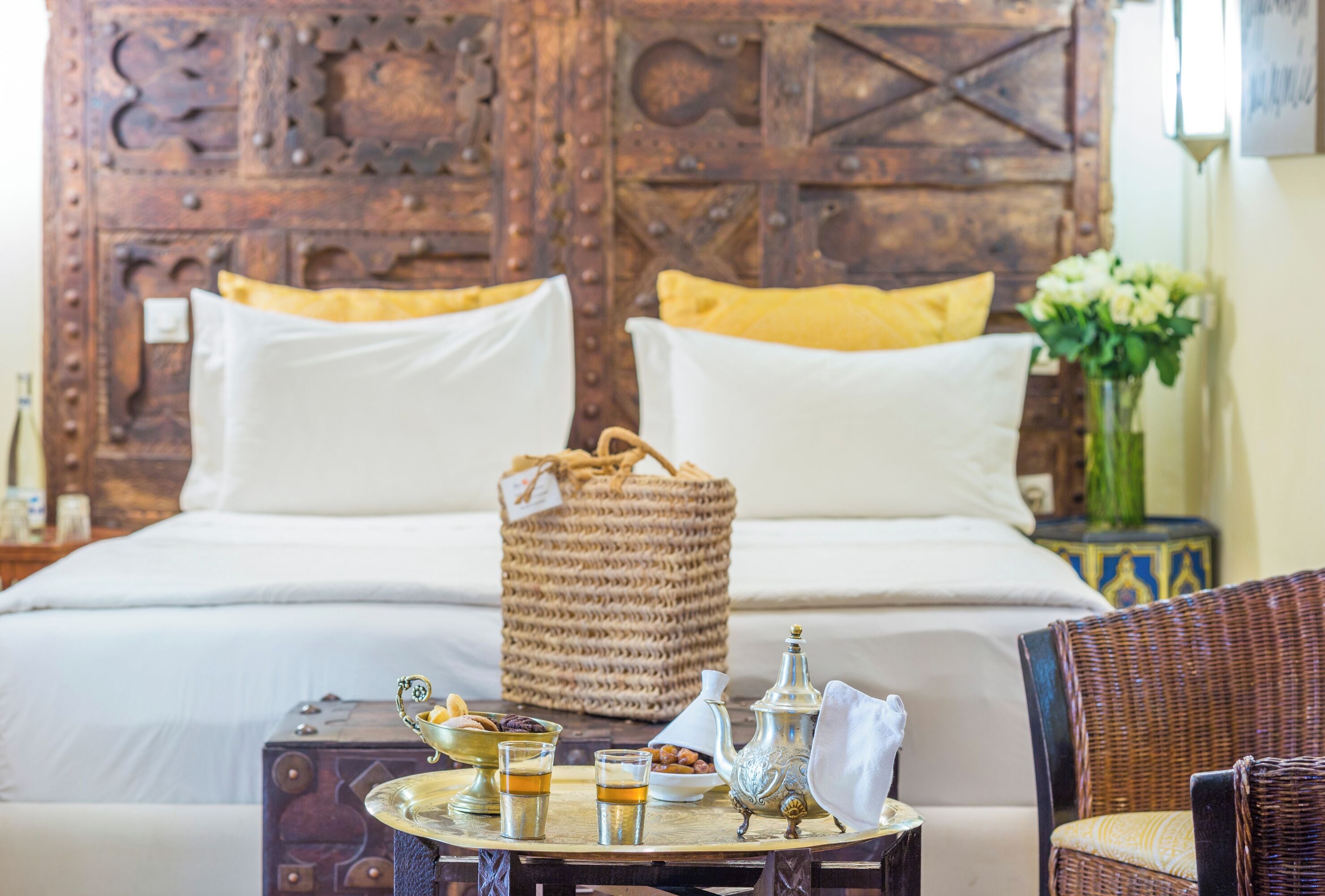 A bed with white and yellow pillows, a woven basket, a tray with a teapot, glasses, and cookies.