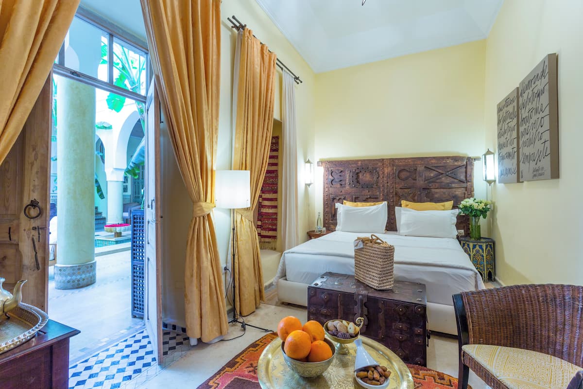 Quarto de hotel com uma cama, uma cadeira de vime, um tabuleiro com frutas e vista para um pátio.