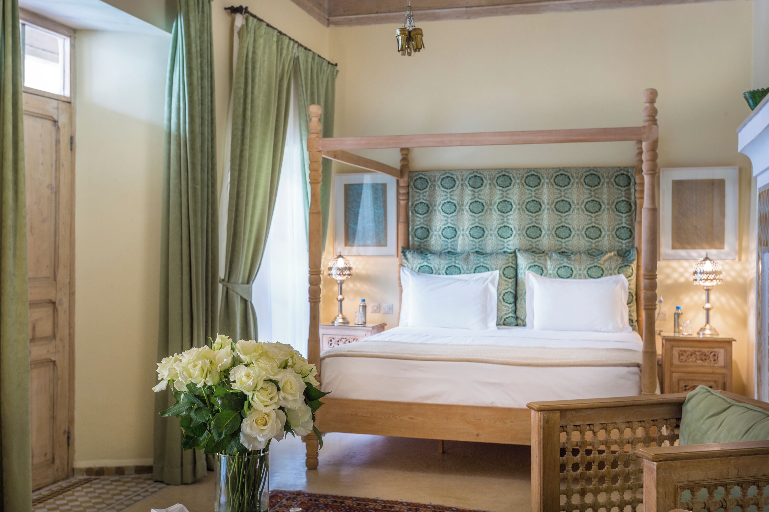 A four-poster bed with a patterned headboard, a bedside table with a lamp, and a vase of flowers on the floor.