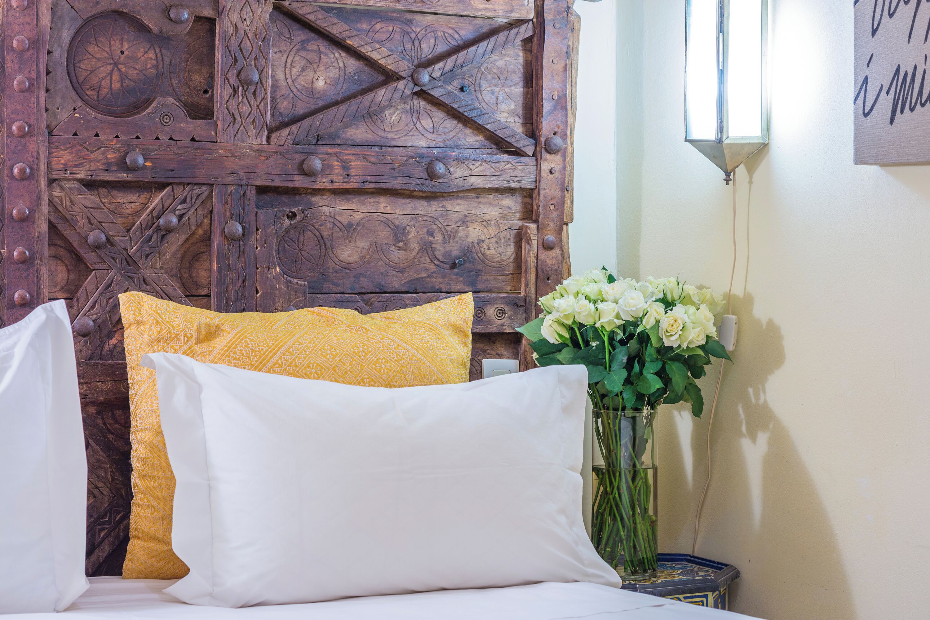 Ein Bett mit weißen und gelben Kissen, ein hölzernes Kopfteil mit aufwendigen Schnitzereien und eine Vase mit weißen Blumen auf einem Nachttisch.