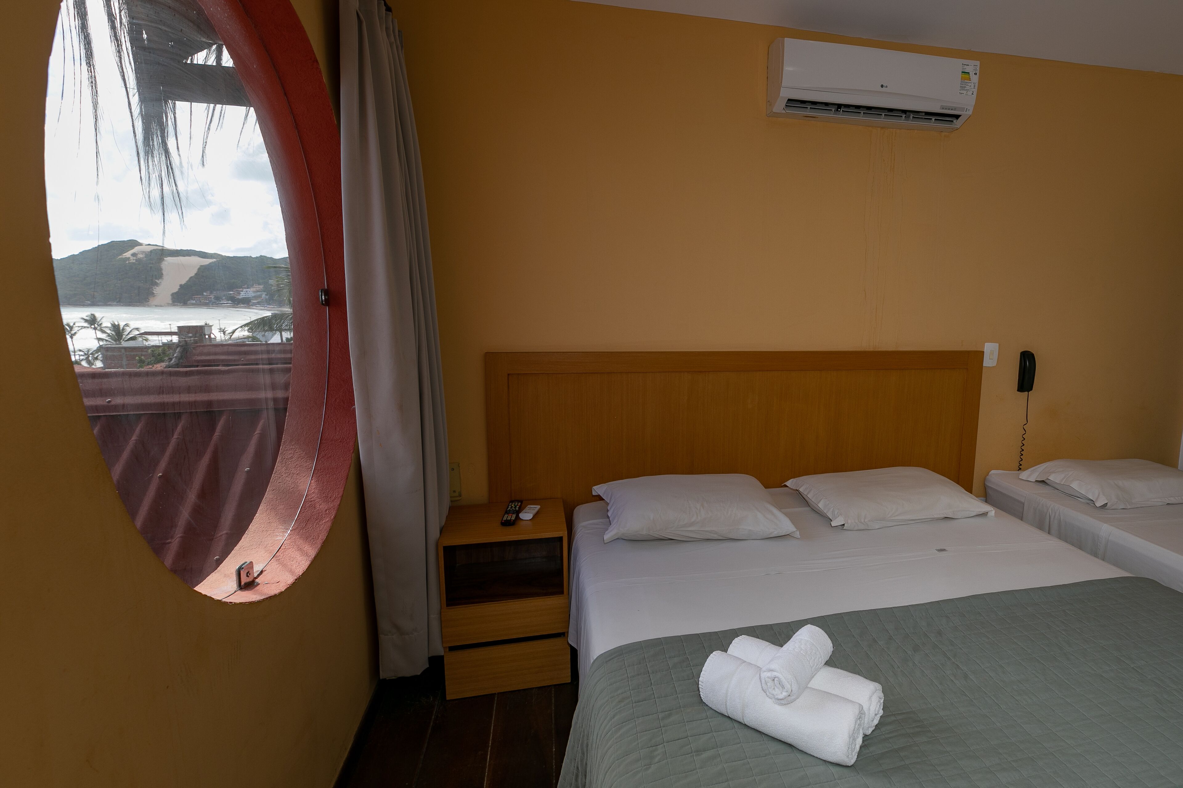 A hotel room with a large window, a bed with white linens, a wooden headboard, a bedside table with a remote control, and an air conditioning unit on the wall.