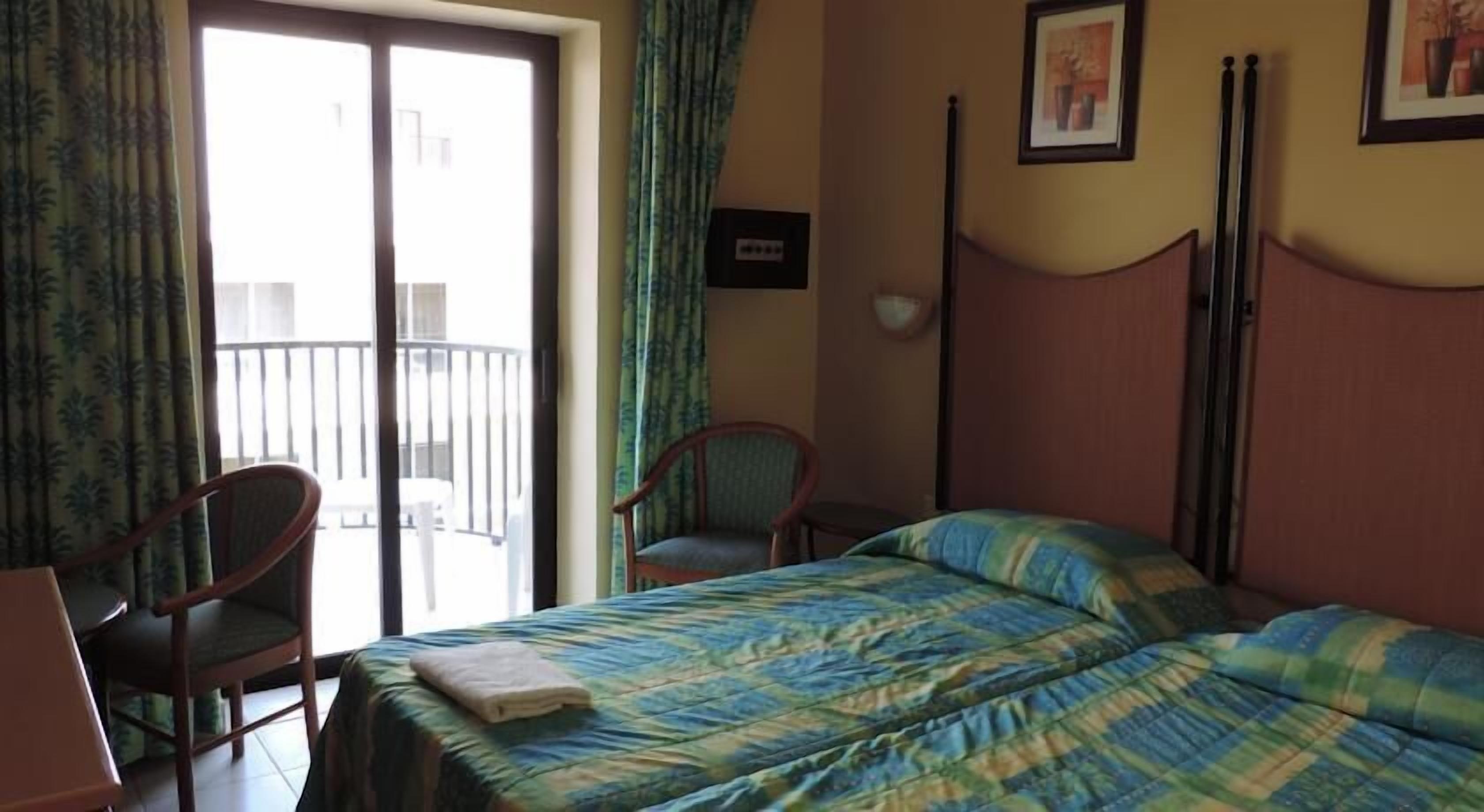 Una habitación de hotel con cama, dos sillas, un escritorio y un balcón con barandilla.