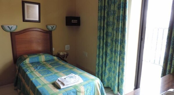 A hotel room with a wooden headboard, a bed with a blue and green checkered bedspread, a folded towel on the bed, a clock on the wall, and a window with curtains. - Euro Club Hotel (St. Paul's Bay)