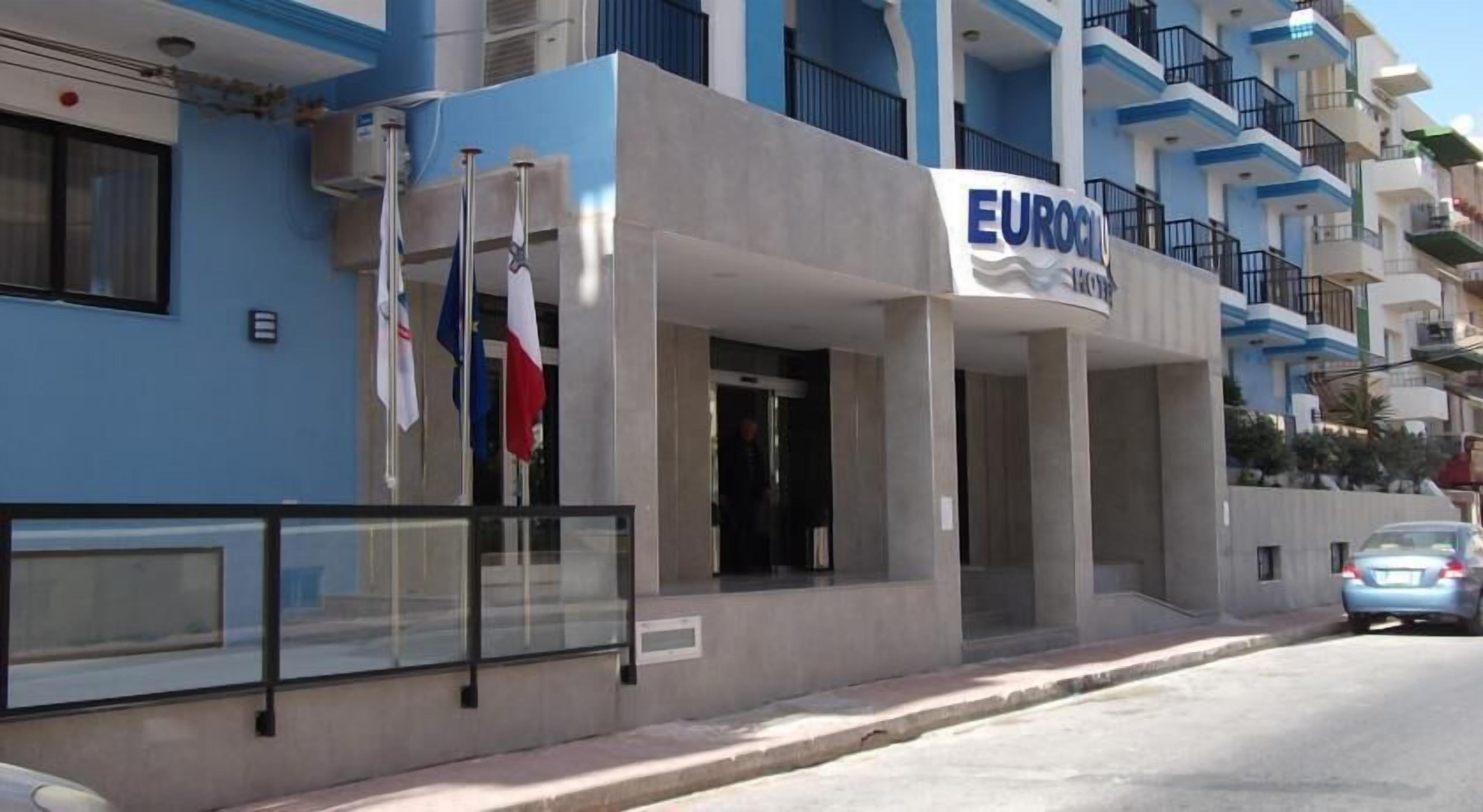 Ein Hotel namens EUROCLIO mit moderner Fassade, Fahnenmasten und einem geparkten Auto davor.
