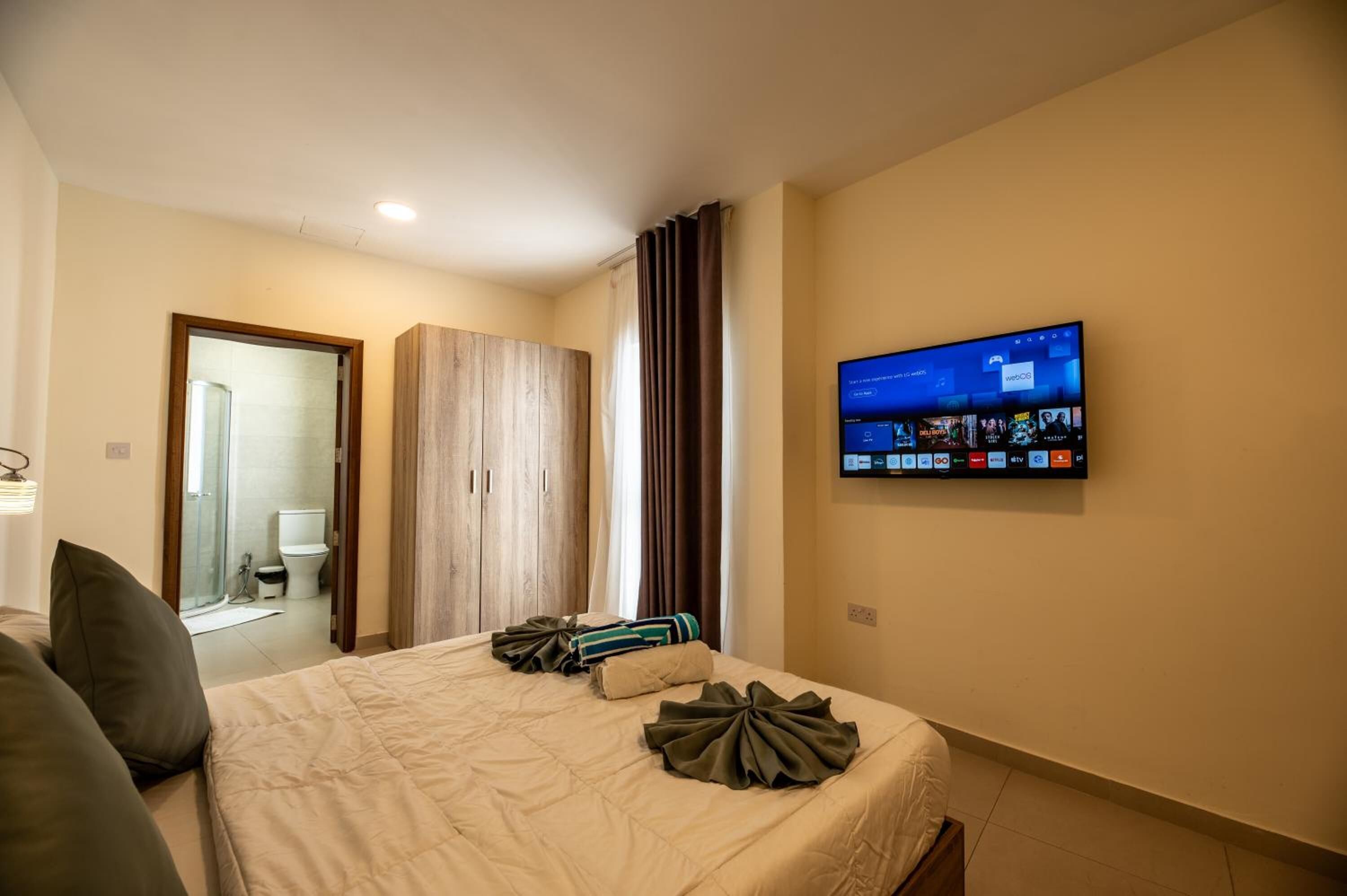 A hotel room with a bed, a TV mounted on the wall, a wooden wardrobe, and a bathroom visible through an open door.