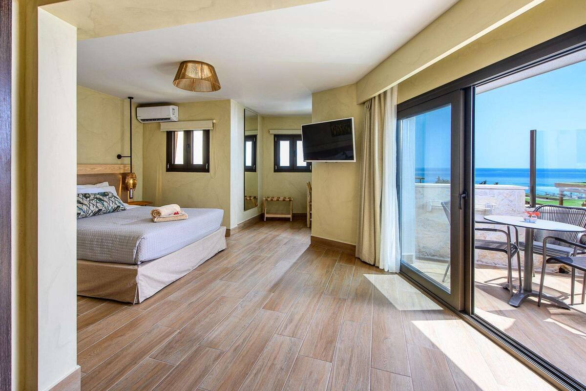 A bedroom with a bed, bedside table, lamp, air conditioning unit, and a TV. There is a balcony with a table and chairs.