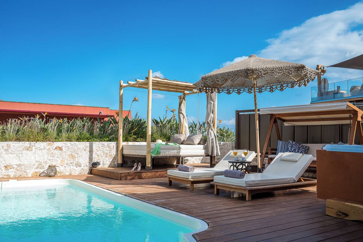 A poolside area with wooden loungers, a pergola with a woven canopy, and a stone wall.