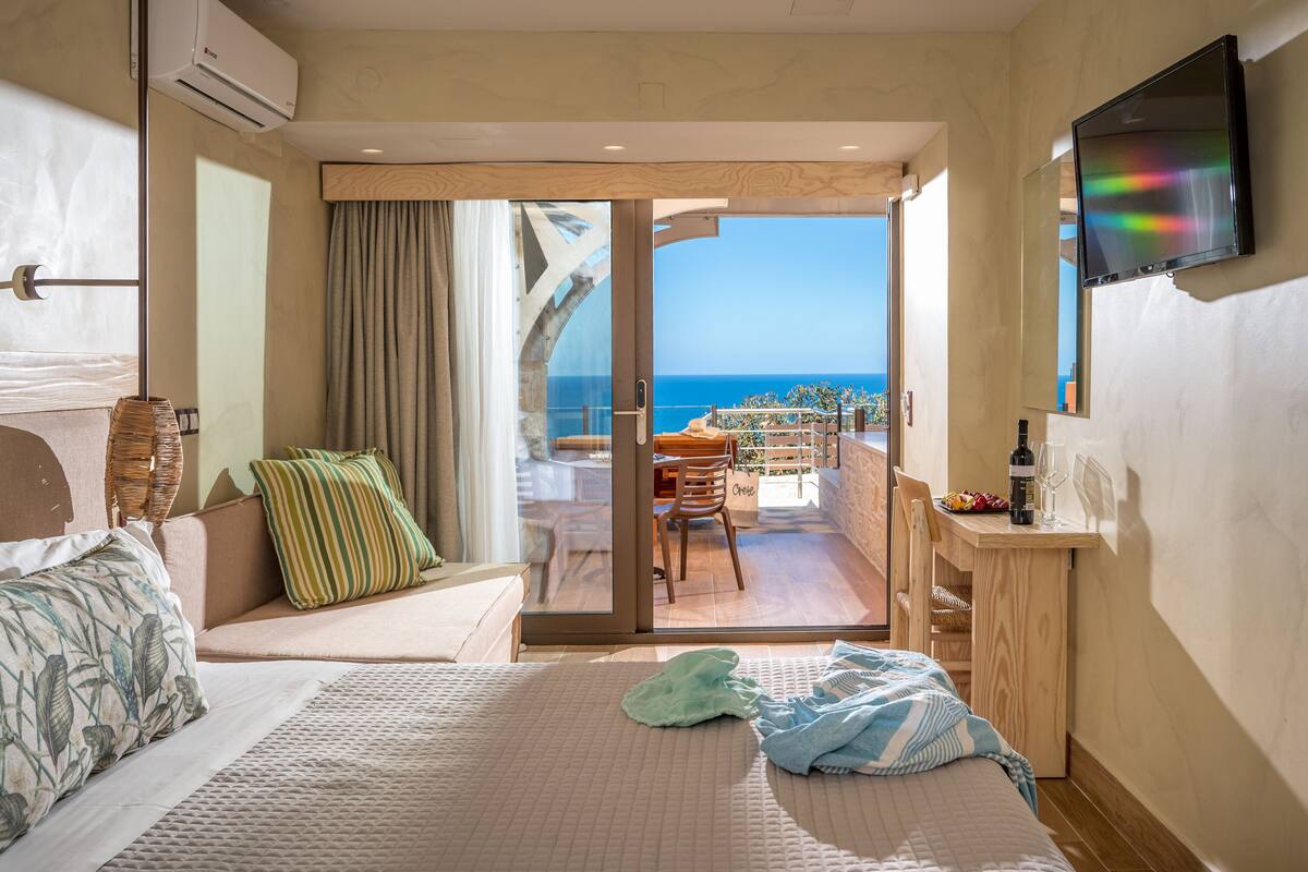 A hotel room with a bed, a seating area with a view of the sea, a TV, and a small table with a bottle and glasses.