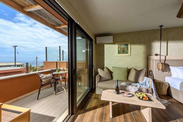 A modern living room with a glass door leading to a balcony, a sofa set, a small table with a bottle and fruit, and a framed artwork on the wall.