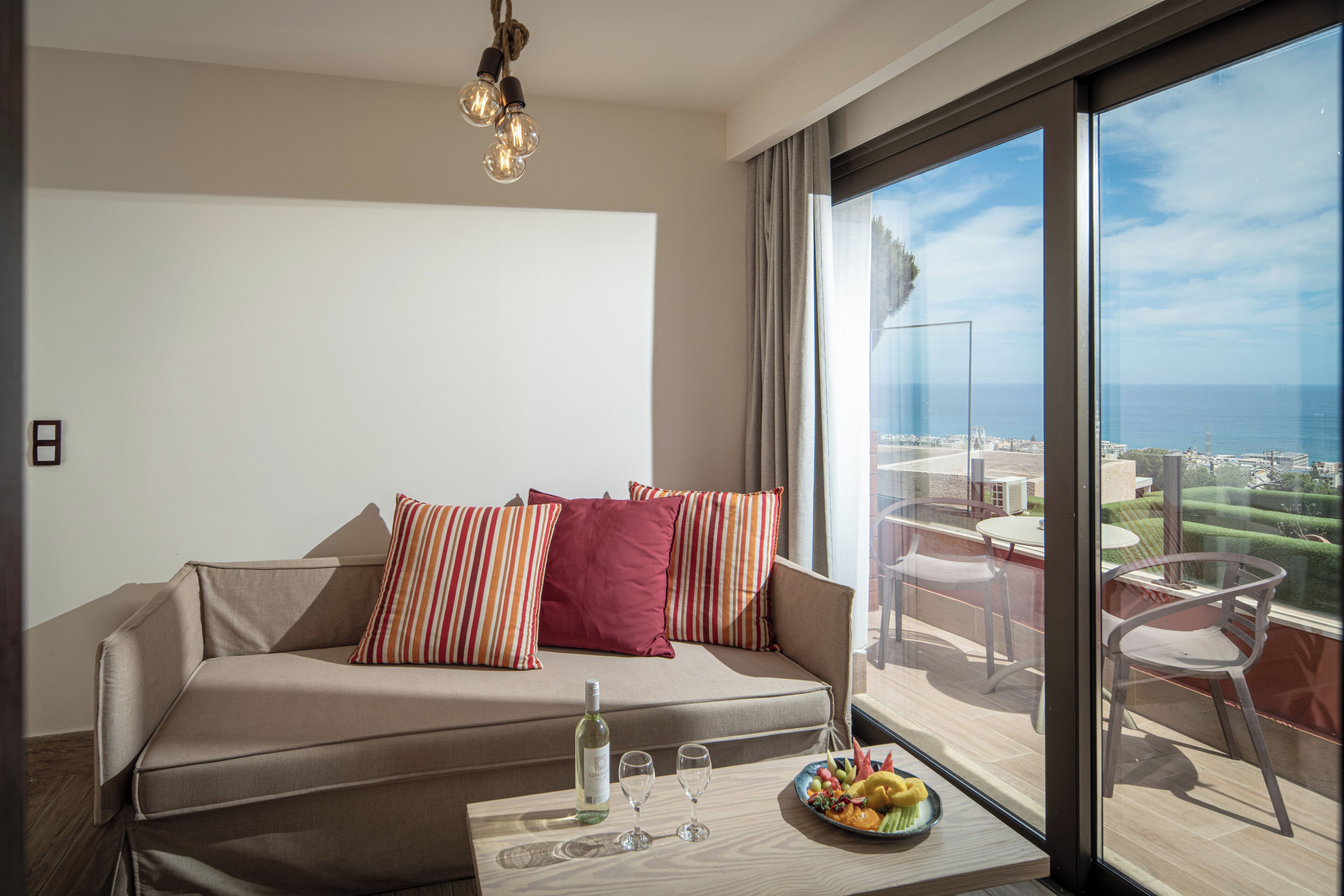 A modern living room with a sofa, a dining table with a bottle and glasses, and a balcony with a view.