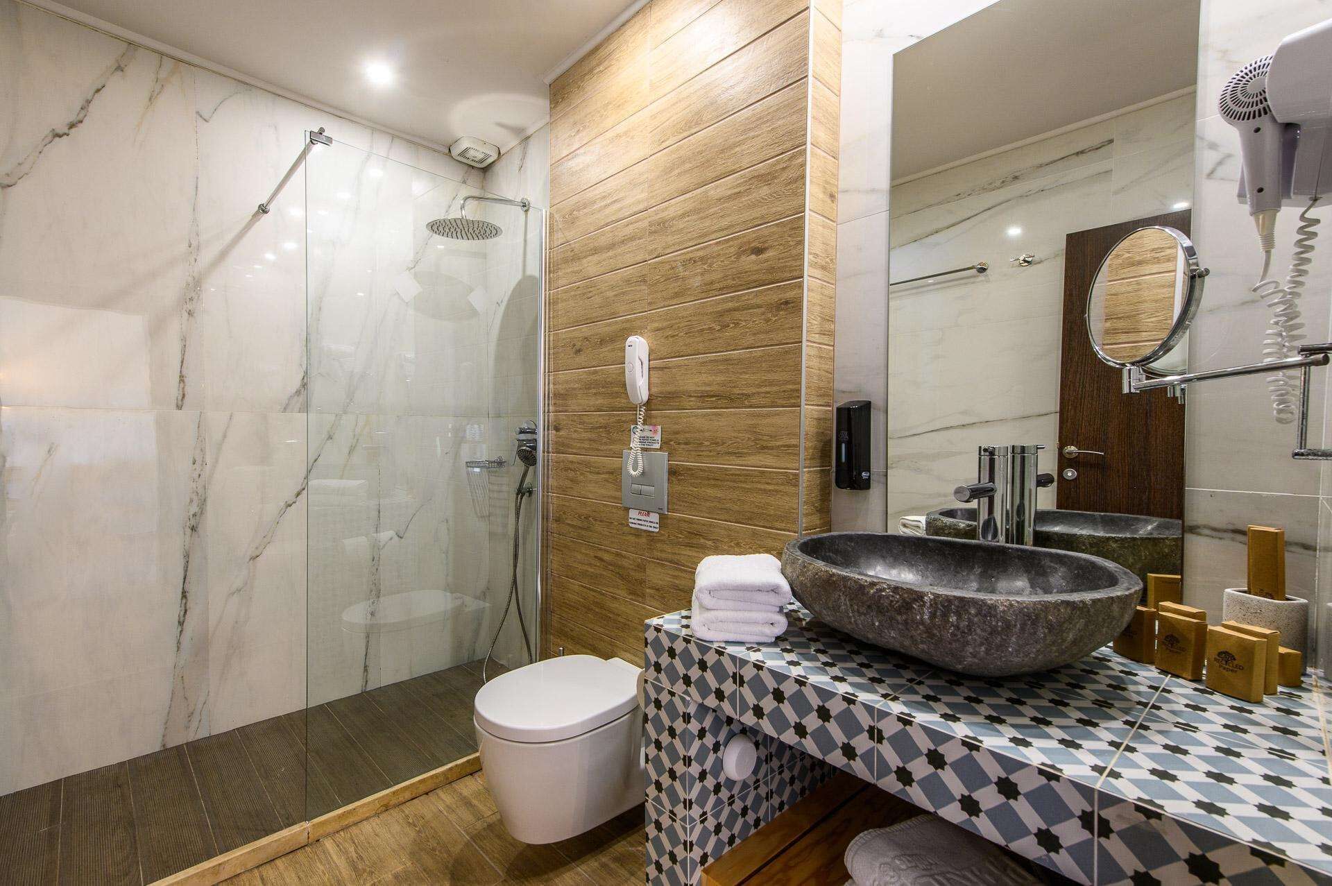 A modern bathroom with a glass-enclosed shower, a marble-tiled wall, a wooden panel accent, a large sink with a stone basin, and a toilet.