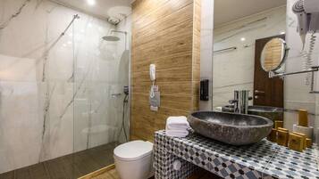 A modern bathroom with a glass-enclosed shower, a marble-tiled wall, a wooden panel accent, a large sink with a stone basin, and a toilet.