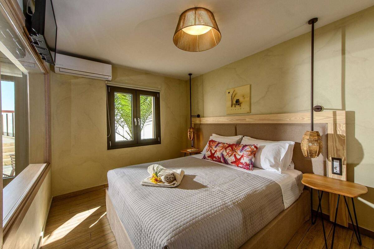 A hotel room with a bed, a bedside table, a lamp, a window with a view of greenery, and an air conditioning unit.