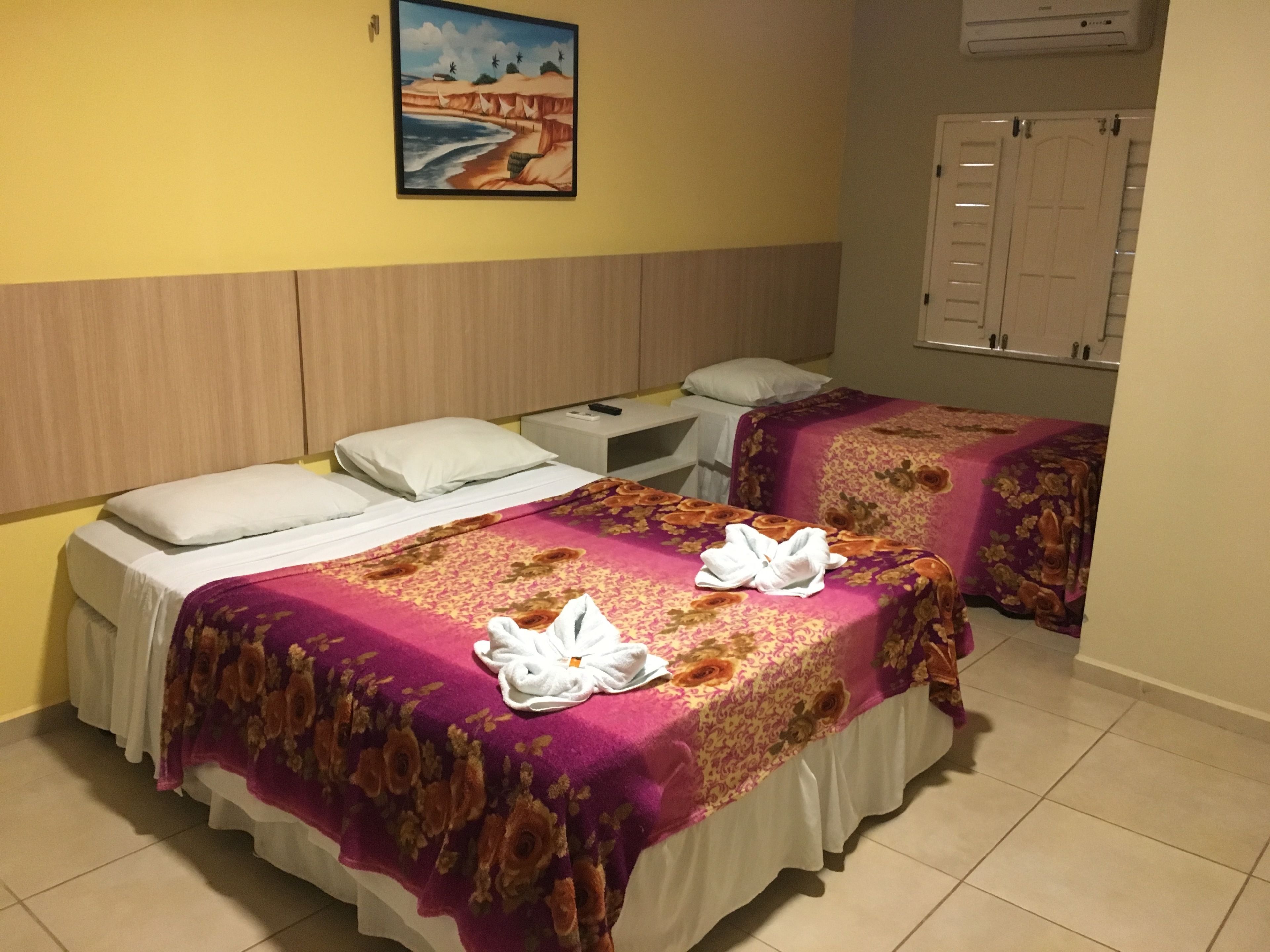 A hotel room with two beds, each with a floral bedspread and white pillows. There is a painting on the wall above the beds, and a white door with a handle on the right side.