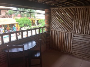 A balcony with a wooden table and chairs, a view of a street with buildings and greenery, and a bamboo fence. - Pousada Latitude (Aracati)