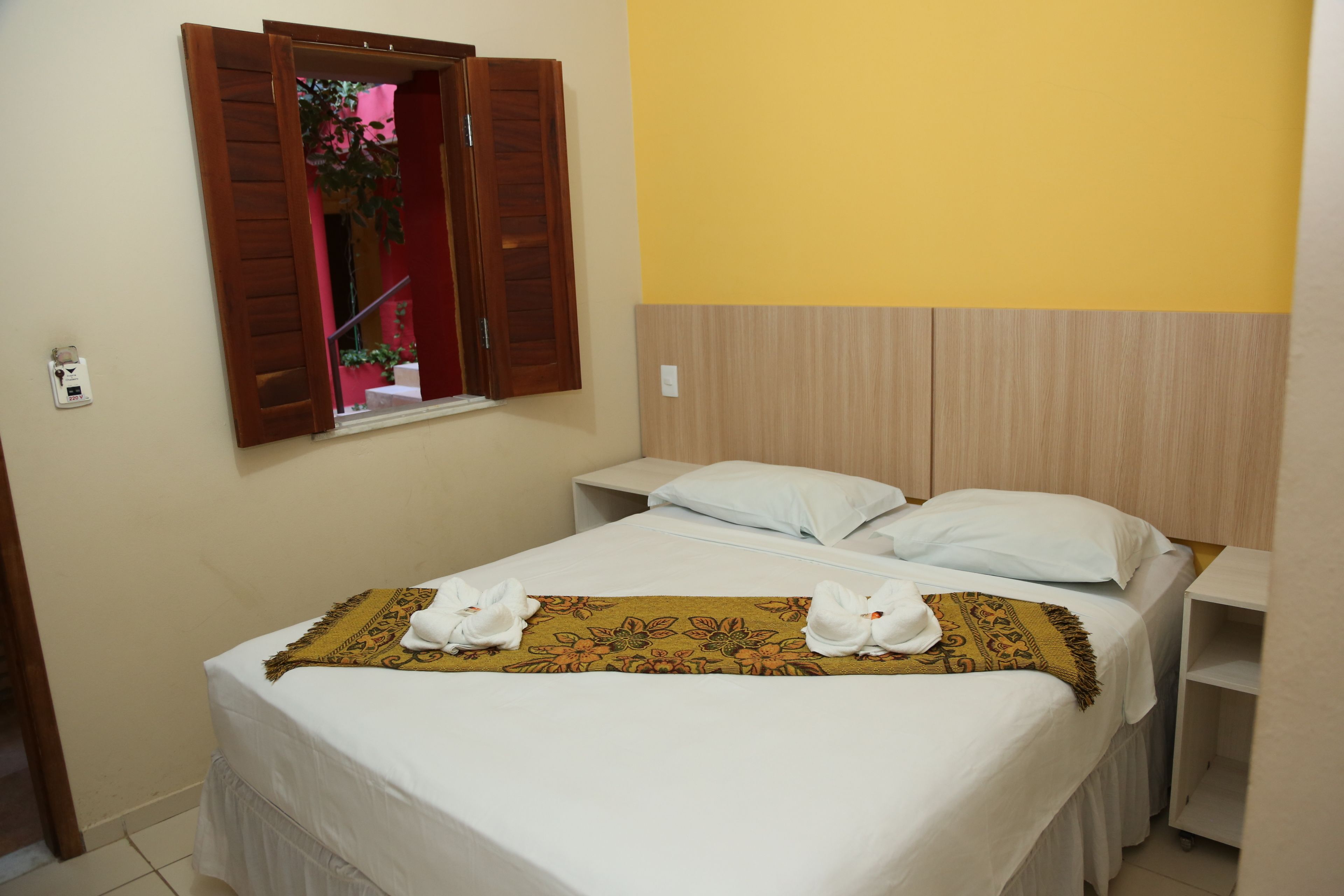 A hotel room with a bed, two pillows, a window with wooden shutters, a bedside table, and a small shelf.