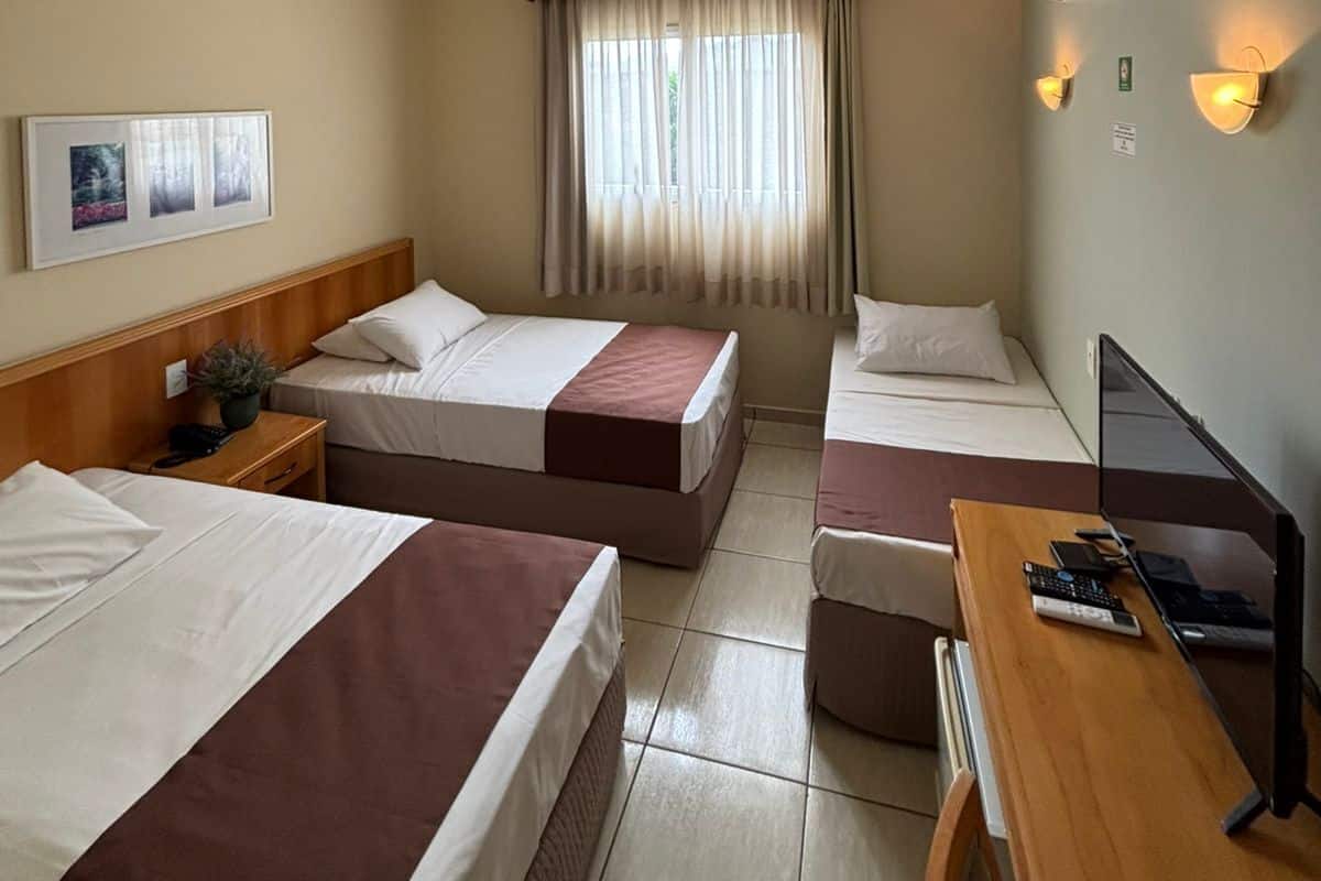 Ein Hotelzimmer mit zwei Betten, einem Schreibtisch mit Computer, einem Fernseher und einem Fenster mit Vorhängen.