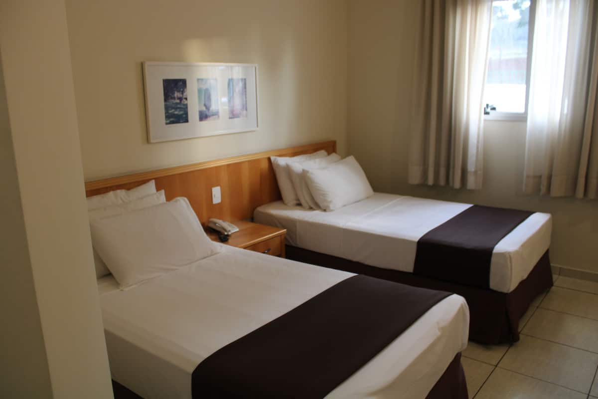 Ein Hotelzimmer mit zwei Betten, einem Holzkopfteil, einem Nachttisch mit Telefon und einem Fenster mit Vorhängen.