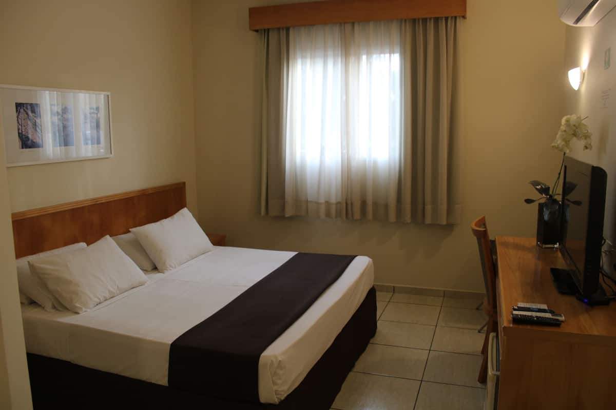 Ein Hotelzimmer mit einem Bett, einem Schreibtisch mit Computer und einem Fenster mit Vorhängen.