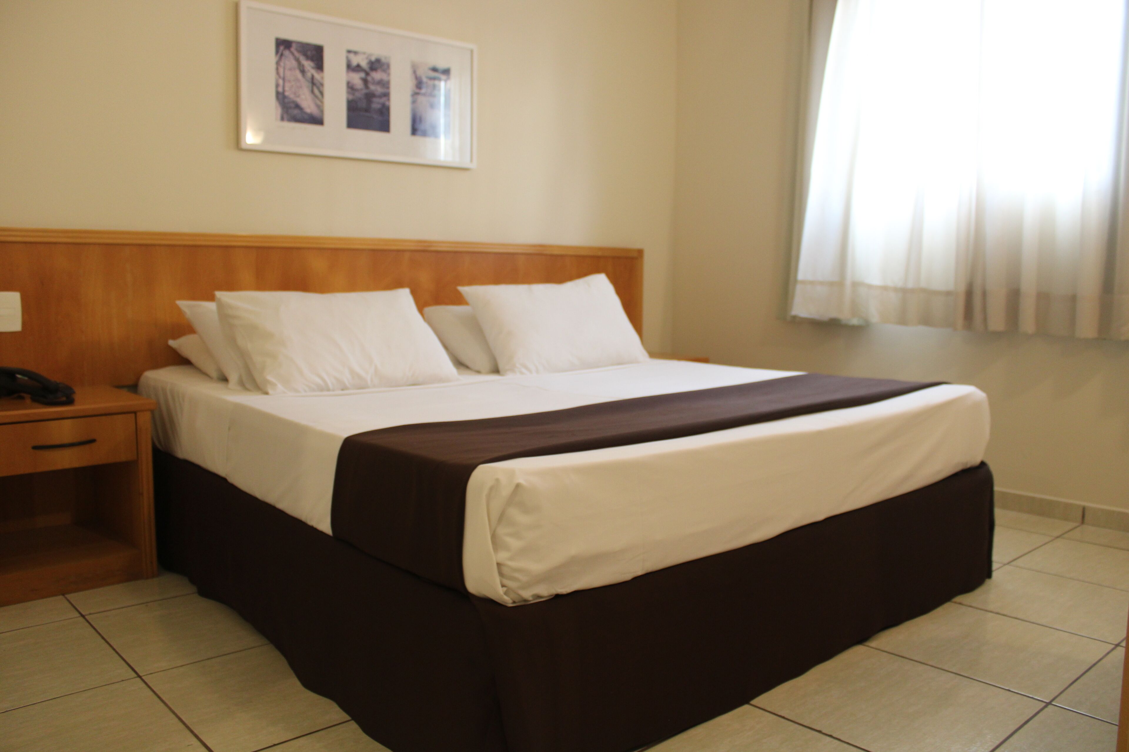 Cama bem arrumada com roupa de cama branca e marrom, cabeceira de madeira e um quadro na parede.