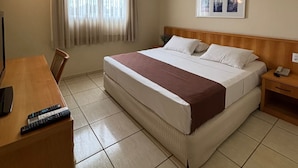 Ein Hotelzimmer mit Bett, Schreibtisch mit Fernbedienung, Nachttisch mit Telefon, Fenster mit Vorhängen und einem gerahmten Bild an der Wand.