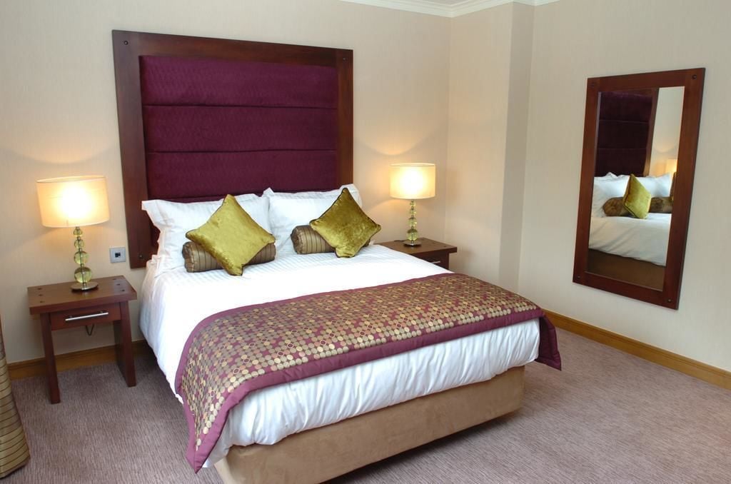 A hotel room with a large bed, two bedside tables with lamps, a large mirror on the wall, and a carpeted floor.
