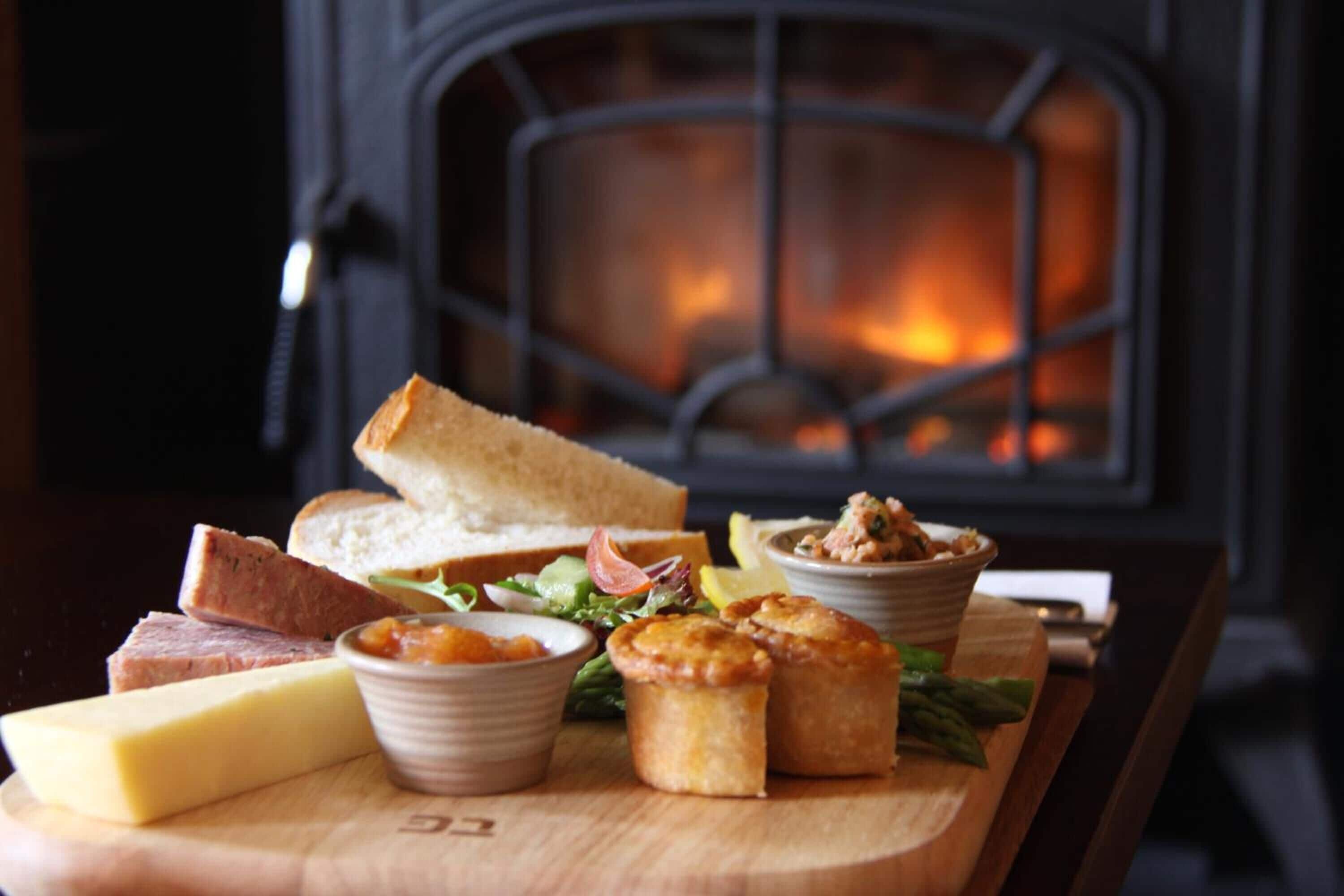 A wooden board with cheese, bread, and small baked goods in front of a fireplace.
