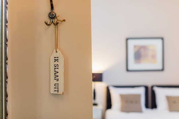 A tag with the text "KIR SLAAP UIT" hanging on a wall, and a blurred background of a room with a bed and pillows.