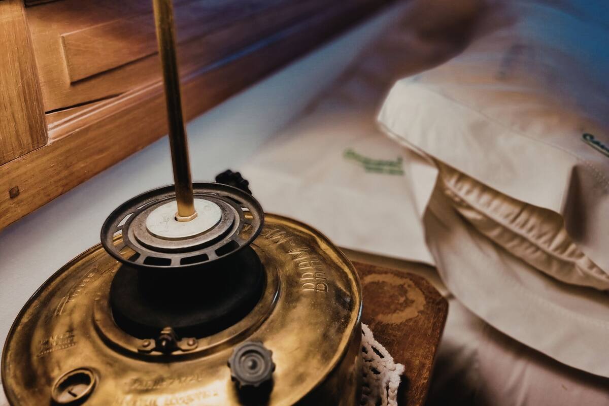 Une lampe en laiton est posée sur une table de chevet en bois, à côté d’un lit agrémenté d’un oreiller blanc et d’une couverture blanche.