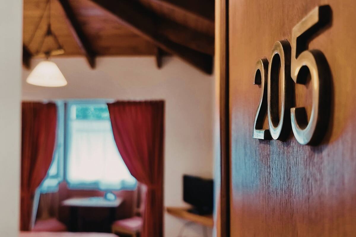 Une chambre d’hôtel avec un plafond en bois, un lit recouvert d’une courtepointe rouge, un bureau avec un ordinateur et une fenêtre ornée de rideaux rouges.