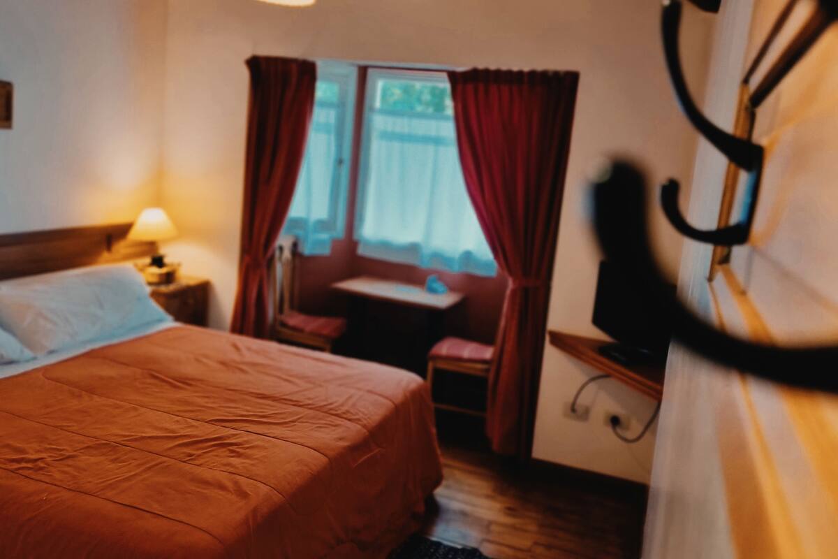 Une chambre à coucher avec un lit, une fenêtre avec des rideaux, une petite table et un téléviseur.