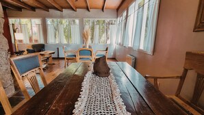 Una mesa de comedor de madera rústica con un camino tejido a ganchillo, rodeada de sillas, en una habitación con ventanas grandes y vigas de madera en el techo.