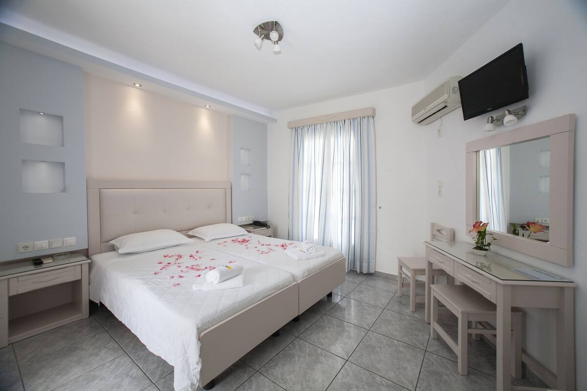 A hotel room with a large bed, a vanity with a mirror, a television mounted on the wall, and a window with curtains.