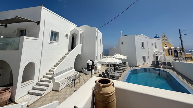 Un espace piscine aménagé sur le toit, avec des bâtiments blancs, un escalier, des chaises longues et un ciel d’un bleu limpide.