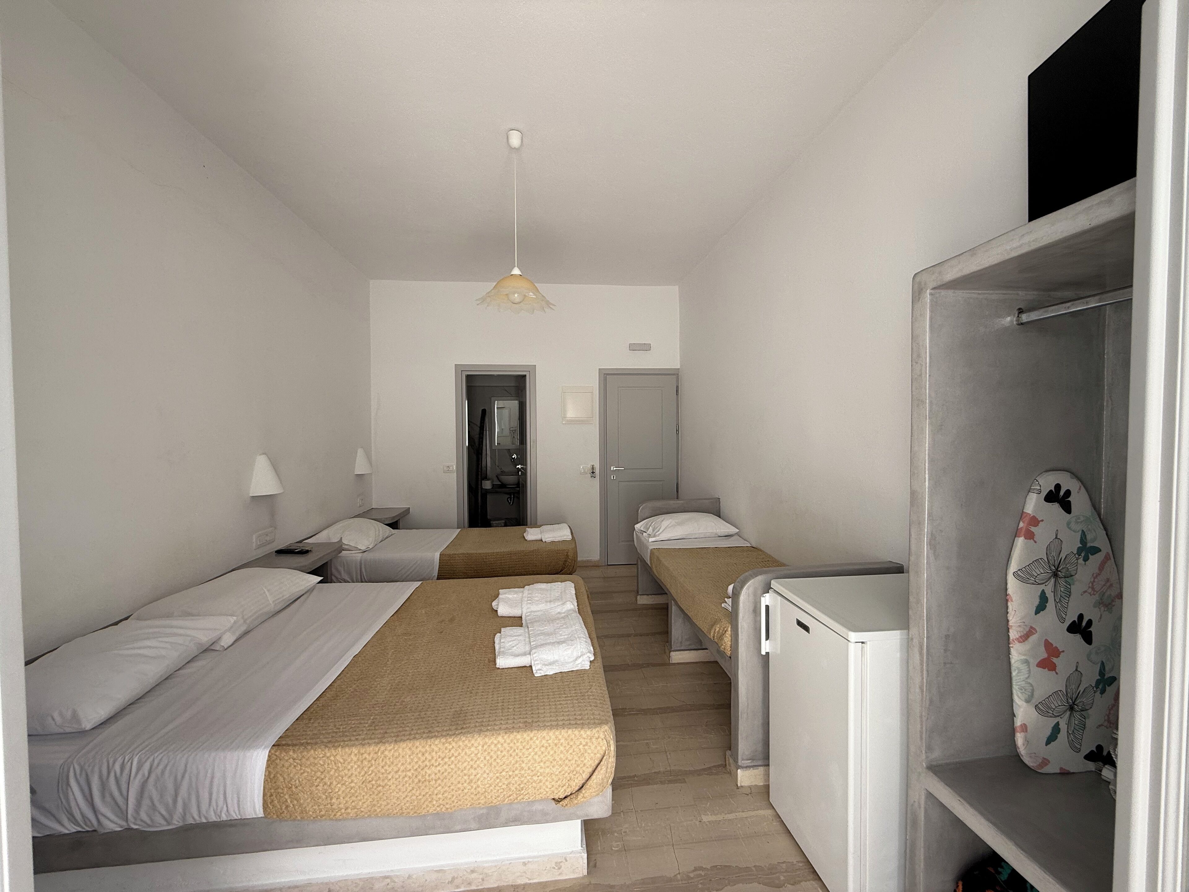 A hotel room with two single beds, a small kitchenette, a wardrobe, and a bathroom visible through an open door.