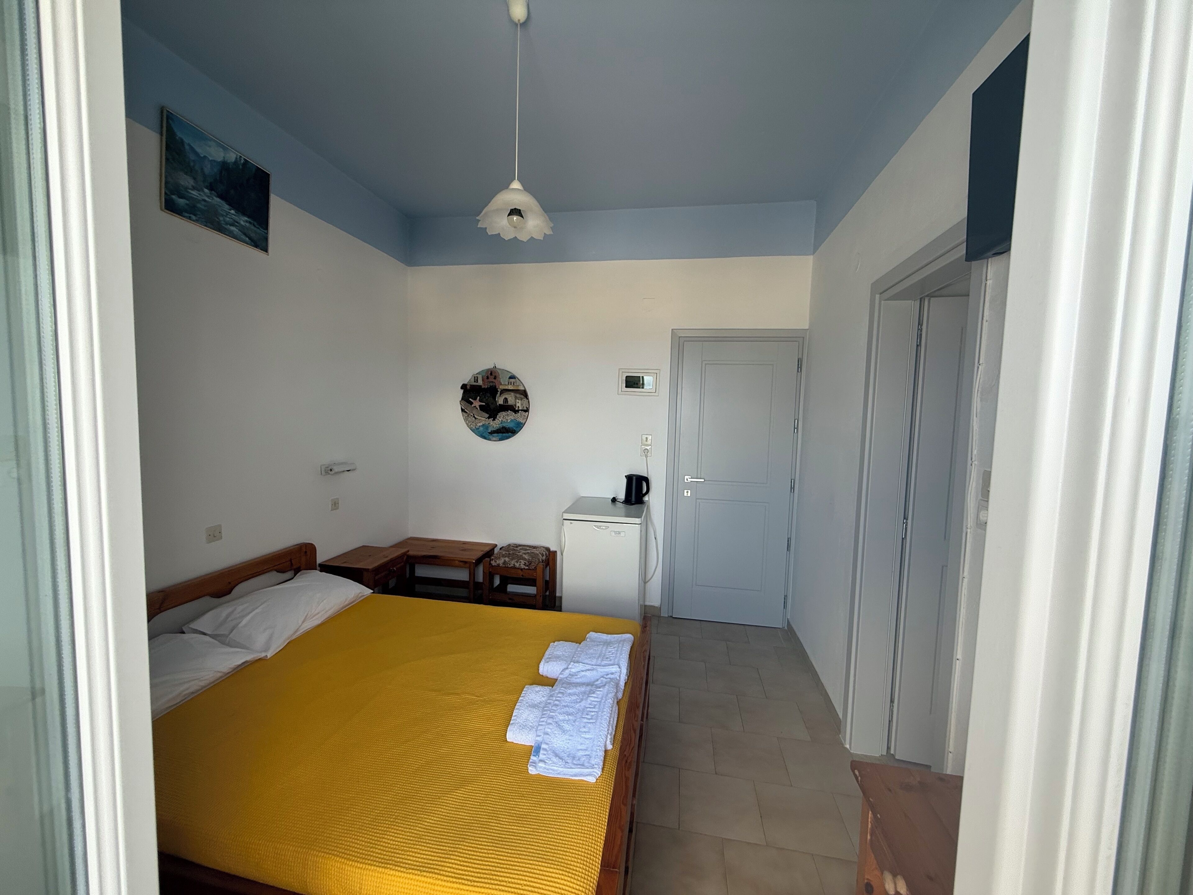 A bedroom with a bed, a desk, a chair, a small refrigerator, and a door.
